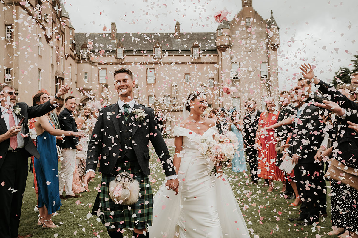 A bride and groom walk towards the camera with a castle behind them as their guests throw pink and white confetti over them