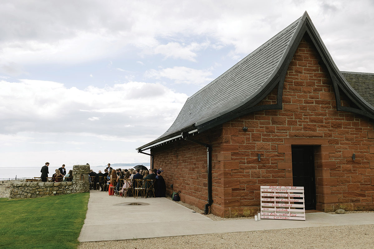 Outdoor wedding ceremony beside a red sandstone boathouse overlooking the sea at a Scottish coastal wedding venue.