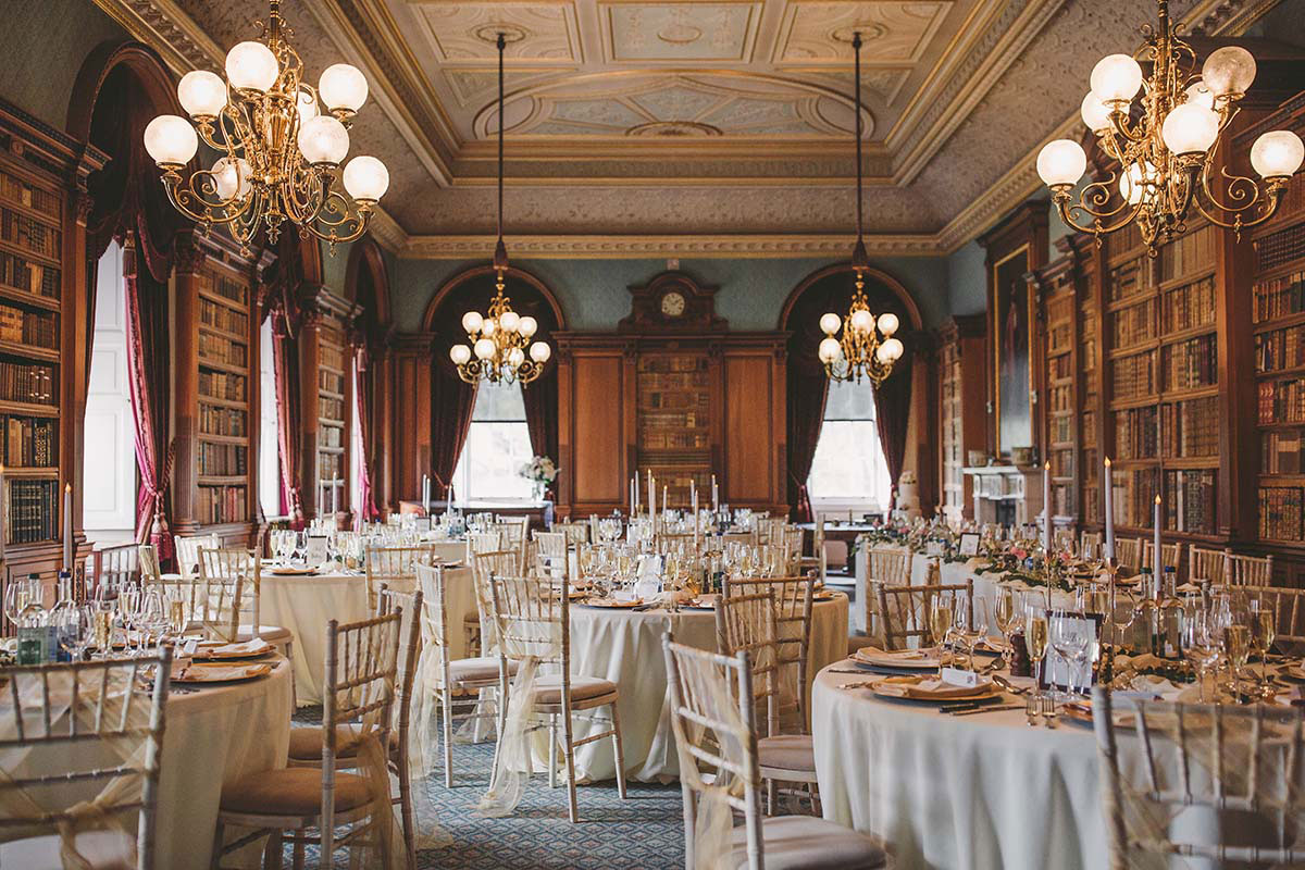 Formal dining room set for a wedding with round tables, gold chairs, chandeliers, and bookshelves lining the walls