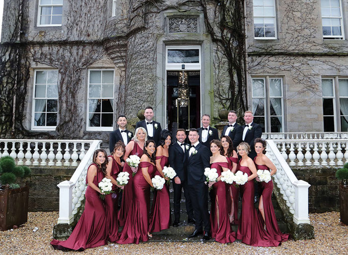 a wedding portrait of two grooms, nine bridesmaids and five men wearing suits on the stone stairs outside Carlowrie Castle