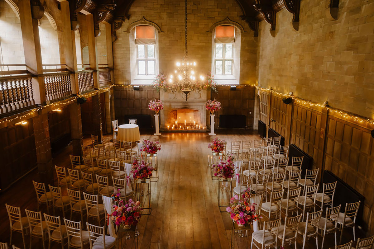 An indoor ceremony setup in a grand wood-panelled ballroom with tall floral arrangements and a lit fireplace.