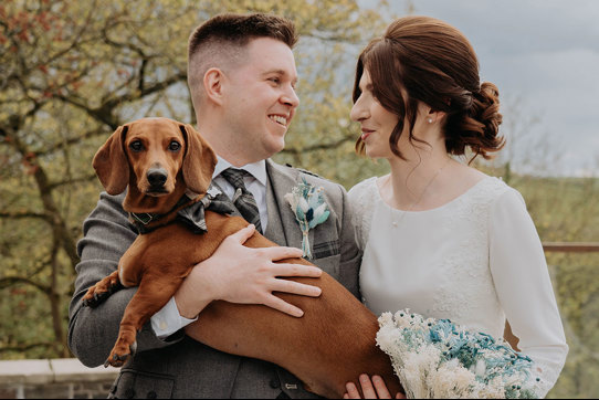  A man in a kilt holds a brown sausage dog who is wearing a bowtie as he stands next to a woman in a white dress