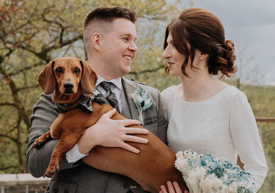  A man in a kilt holds a brown sausage dog who is wearing a bowtie as he stands next to a woman in a white dress