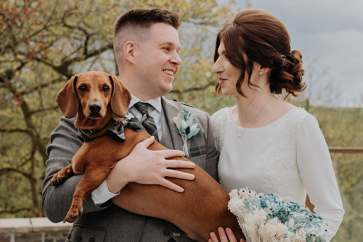 A man in a kilt holds a brown sausage dog who is wearing a bowtie as he stands next to a woman in a white dress