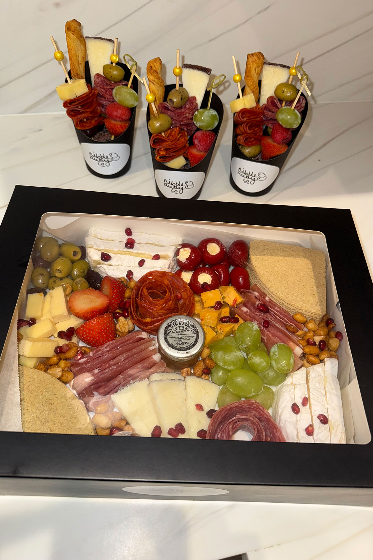 A boxed grazing platter filled with charcuterie, cheeses, olives, grapes, strawberries, stuffed peppers, oatcakes and nuts, with three individual graze cups displayed behind it