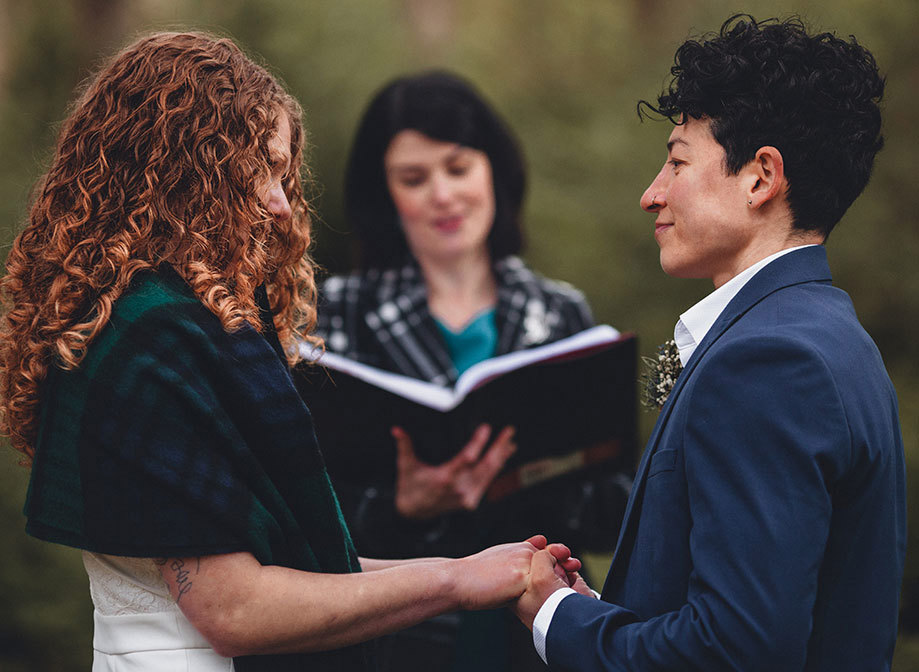 Wedding ceremony in a forest between two women with a female celebrant holding a book 
