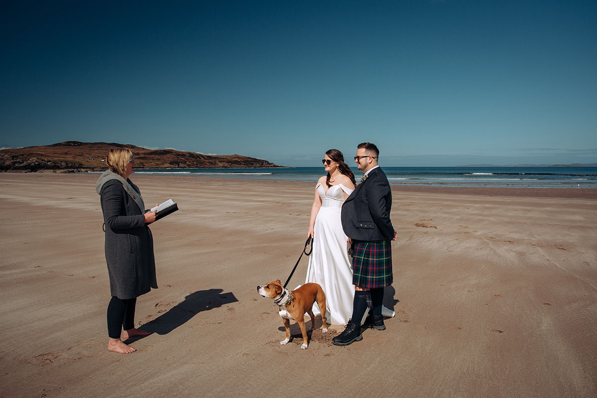 Bride and groom stand barefoot on Achnahaird Bay beach with their celebrant and dog during a relaxed Scottish elopement ceremony.