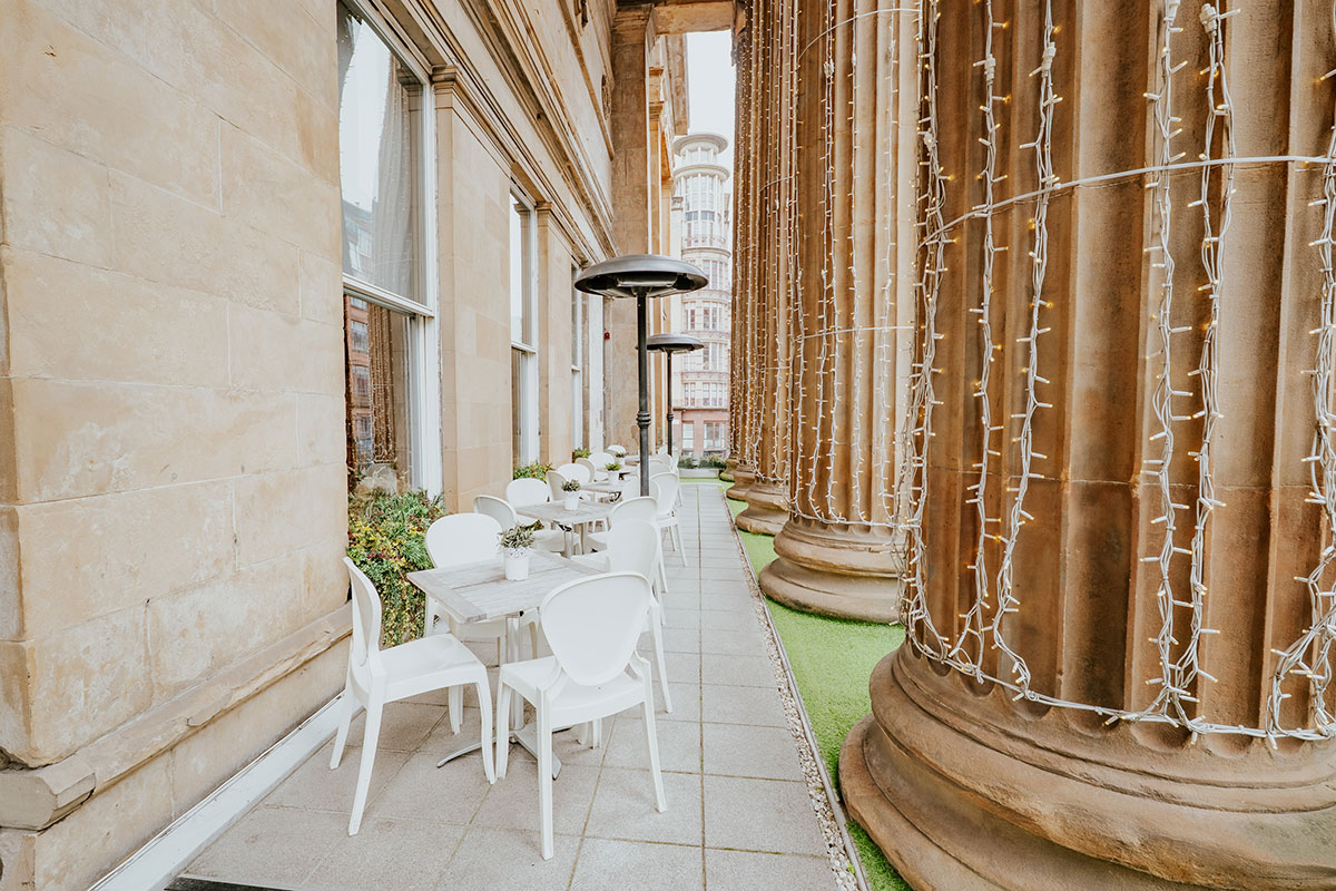 A narrow outdoor terrace with café seating set between the venue’s tall stone columns.