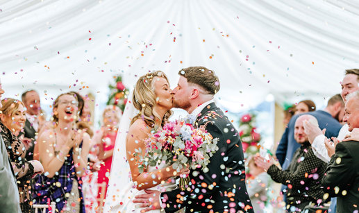 bride and groom kiss while being showered in multicoloured confetti as guests clap in background