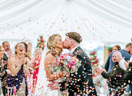 bride and groom kiss while being showered in multicoloured confetti as guests clap in background