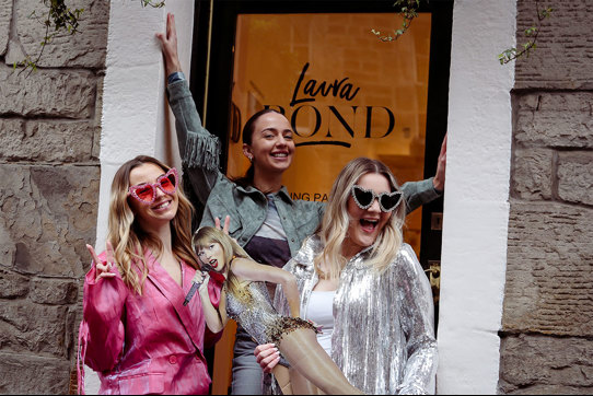 Three women stand in front of a glass door with the words 'Laura Bond' written on it, wearing heart shaped sunglasses and holding a Taylor Swift cardboard cutout
