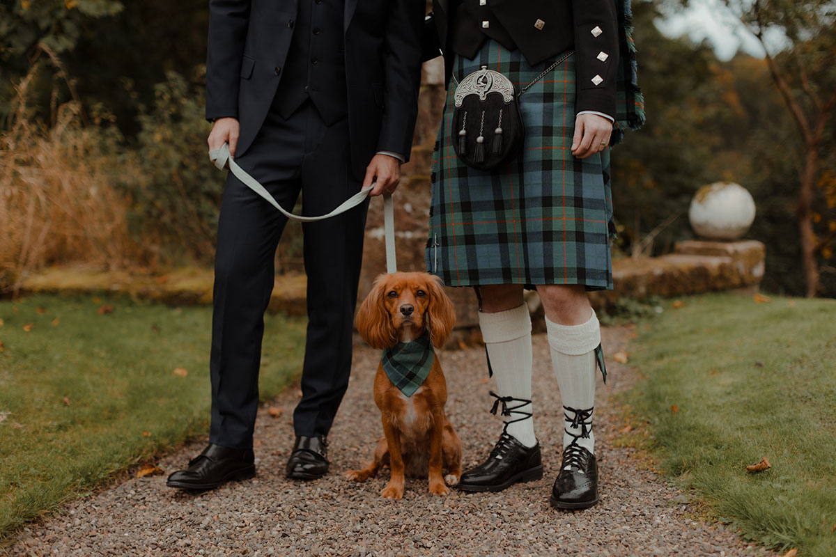 Lower view of two grooms—one in a suit and one in a Gunn tartan kilt—standing with their dog on the lead during portraits at Newhall Estate