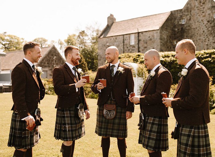 five men wearing brown kilt outfits hold brown hipflasks in a garden setting with stone buildings in the background