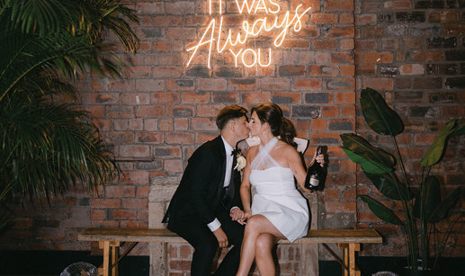 bride and groom sitting on a bench kissing at Engine Works. They are in front of a brick wall below a neon sign.