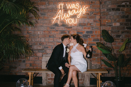 bride and groom sitting on a bench kissing at Engine Works. They are in front of a brick wall below a neon sign.