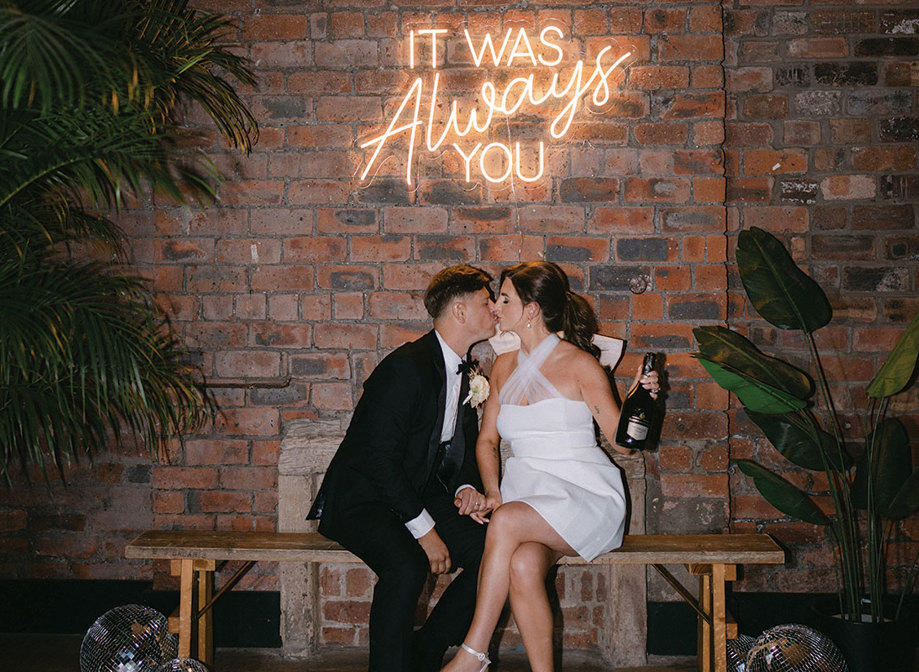 bride and groom sitting on a bench kissing at Engine Works. They are in front of a brick wall below a neon sign.