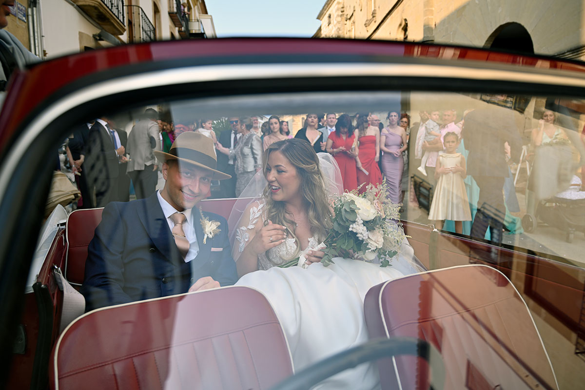 view of bride in white wedding dress and groom in fedora and blue suit through the front window of a red convertible car
