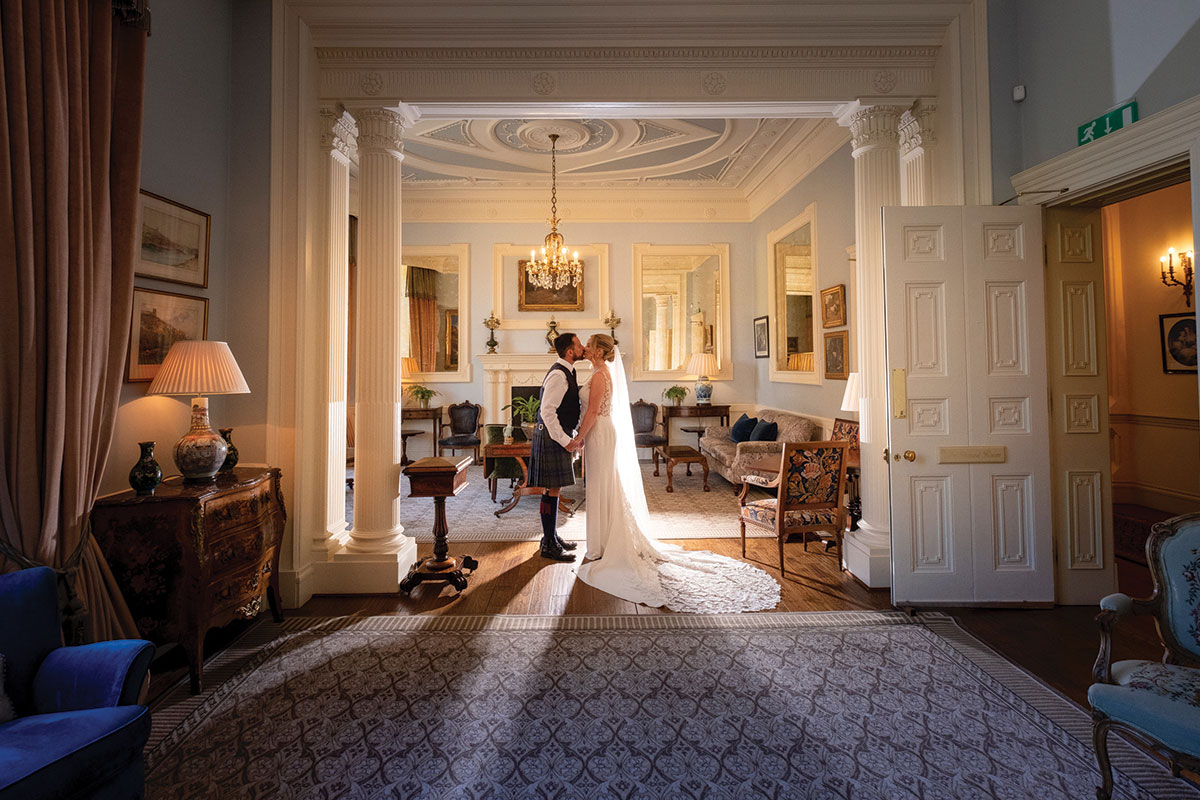 Bride and groom standing beneath grand pillars inside Crossbasket Castle, an elegant Scottish castle wedding venue with chandeliers and historic interiors.