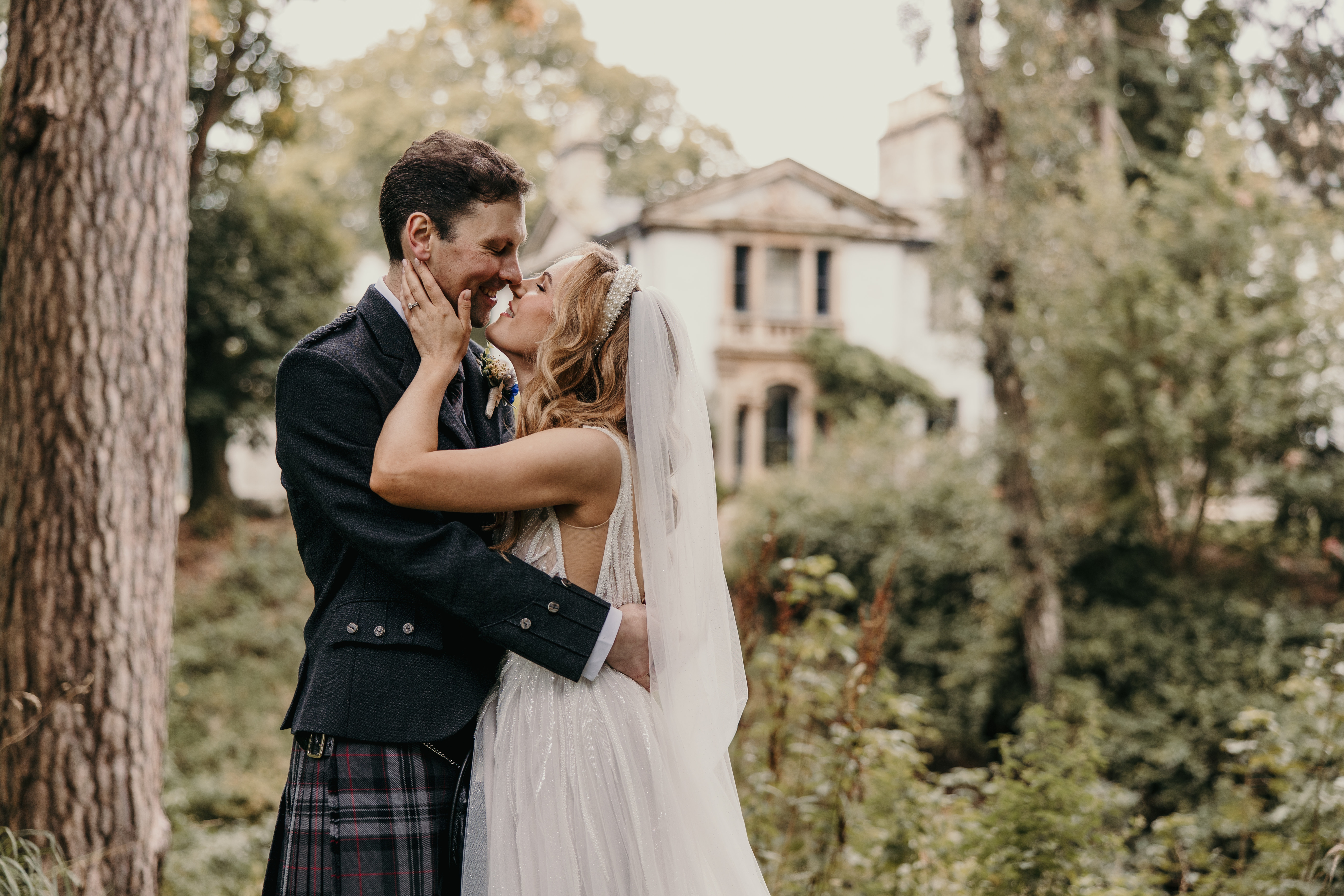 bride holds hands up to face of groom in a kilt and they touch noses stood outside by trees and other greenery with a faded white building in the background