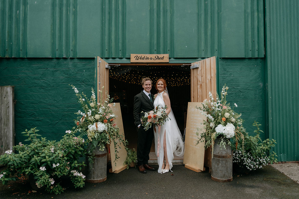 Bride and groom standing outside rustic barn entrance decorated with floral installations
