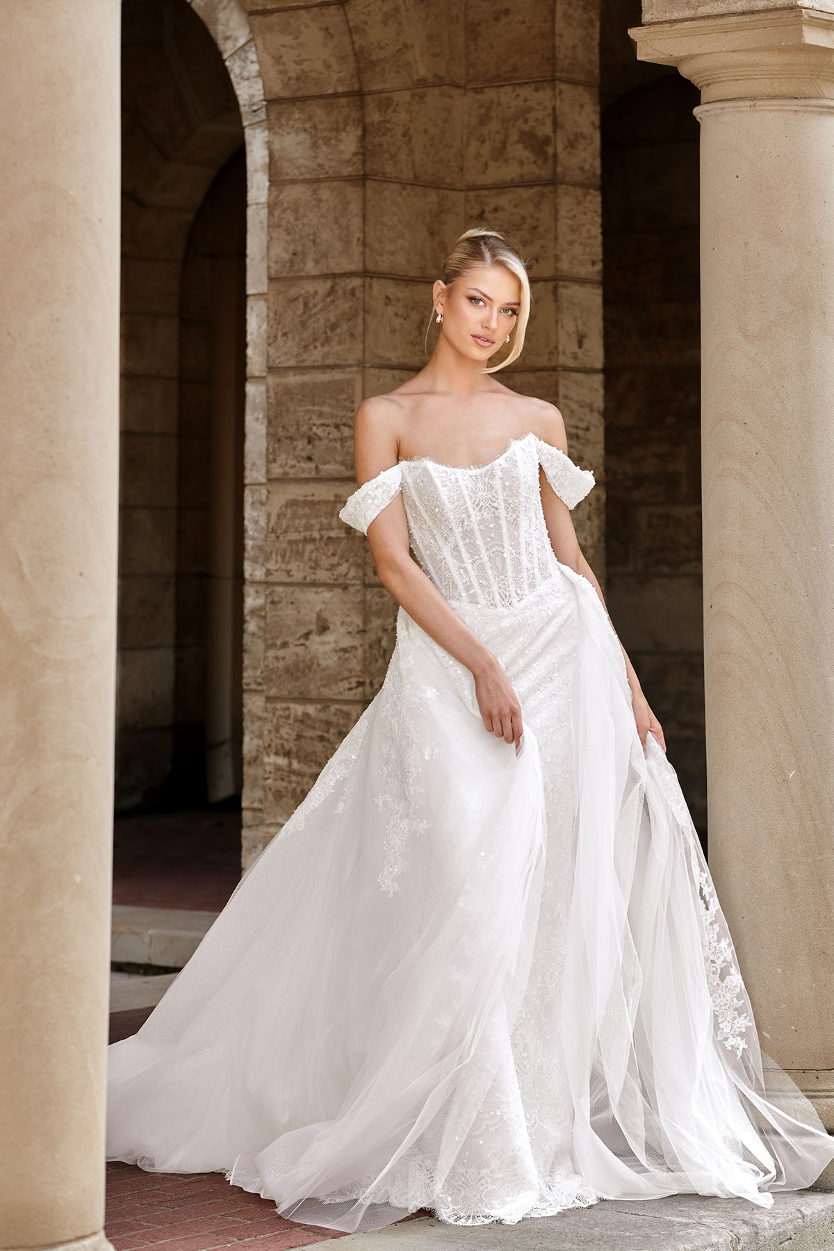 Bride in an off-the-shoulder corset wedding dress with fitted bodice, subtle beading and a flowing tulle skirt, posed between stone columns