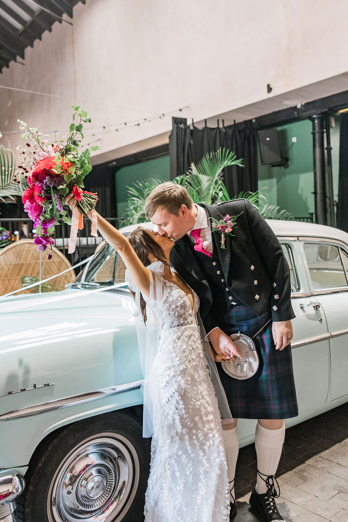 Bride and groom kissing beside a vintage wedding car while holding a colourful bouquet