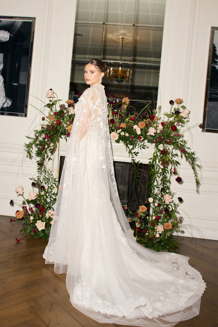 brunette bride with her hair tied back wears floral embroidered wedding dress covered by long matching cape as she stands in front of fireplace decorated with pink and red roses and lots of greenery