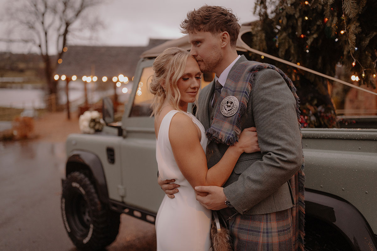 blonde woman in sleeveless white dress holds on to man in in green jacket and red, blue, green kilt with matching fly plaid, both stood in front of green truck with Christmas tree in the back