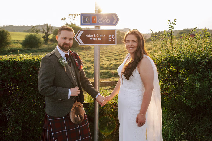A bride and groom stand in front of a sign that says 'Helen & Grant's Wedding' in front of a field