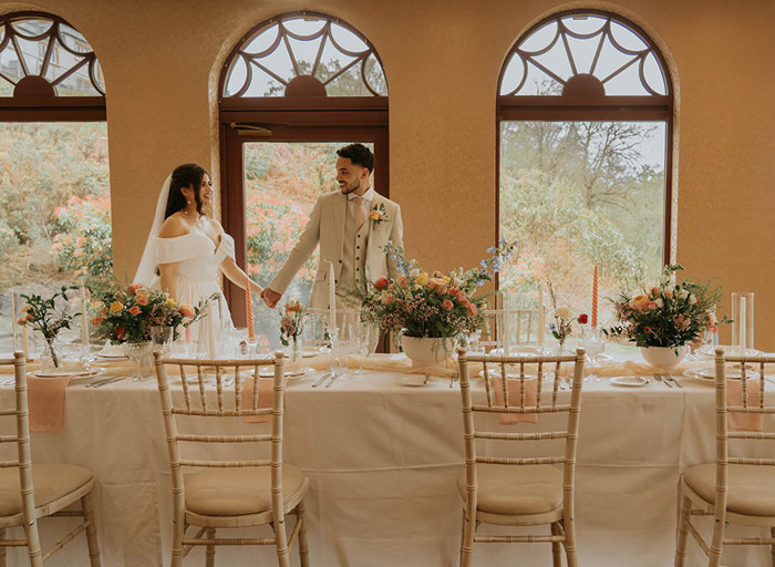 A bride and groom hold hands as they walk past a long table set with plates, glasses and flowers