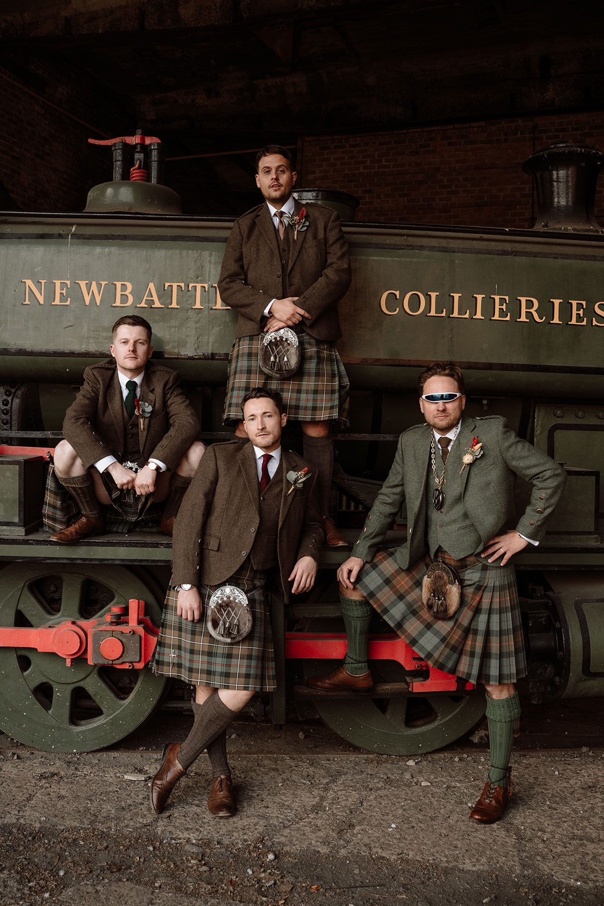 Groomsmen in matching tartan kilts pose in front of a historic steam locomotive, creating a striking industrial wedding portrait