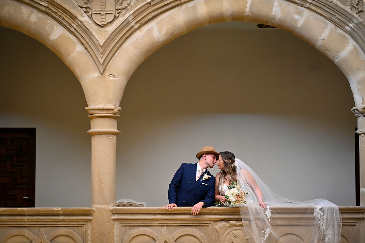 bride with long trailing veil stands by balcony, leaning to the side to kiss groom in dark blue suit and a brown fedora