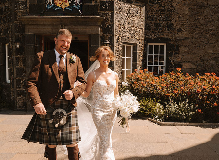a bride wearing an ornate lace wedding dress with sweetheart neckline and spaghetti straps walks arm in arm with a man wearing a brown kilt. They are outside a stone building and there are bushes with orange flowers in the background