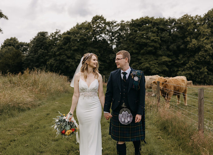 A bride and groom look at each other as they walk towards the camera along a verge of grass with a Highland Cow in a filed behind them