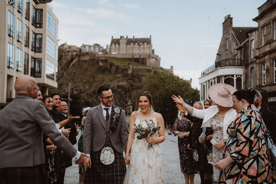 Bride and groom walk through confetti shower with view of Edinburgh Castle in background 