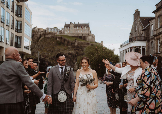 Bride and groom walk through confetti shower with view of Edinburgh Castle in background 
