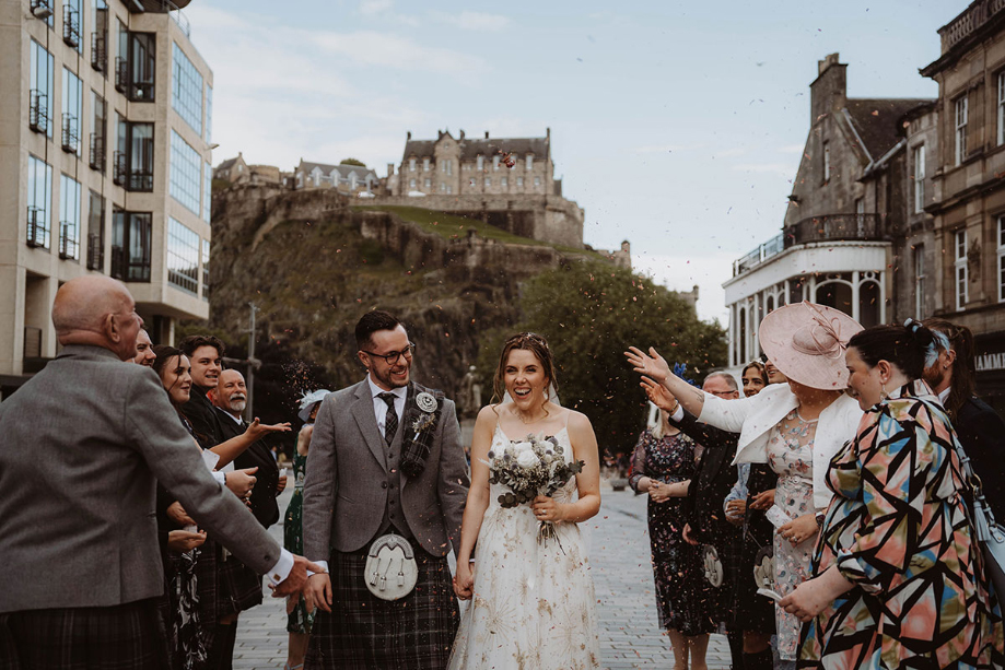 Bride and groom walk through confetti shower with view of Edinburgh Castle in background