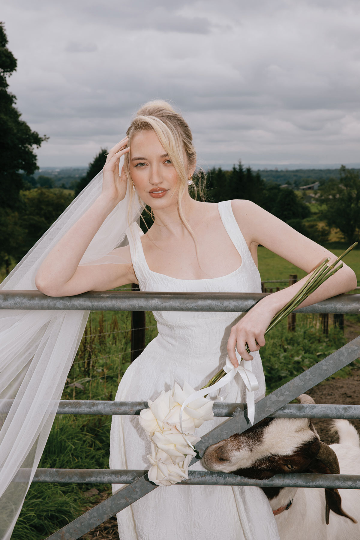 A bride in a square-neck white gown leans on a farm gate with a bouquet of white flowers, while a goat reaches through the gate towards the bouquet.