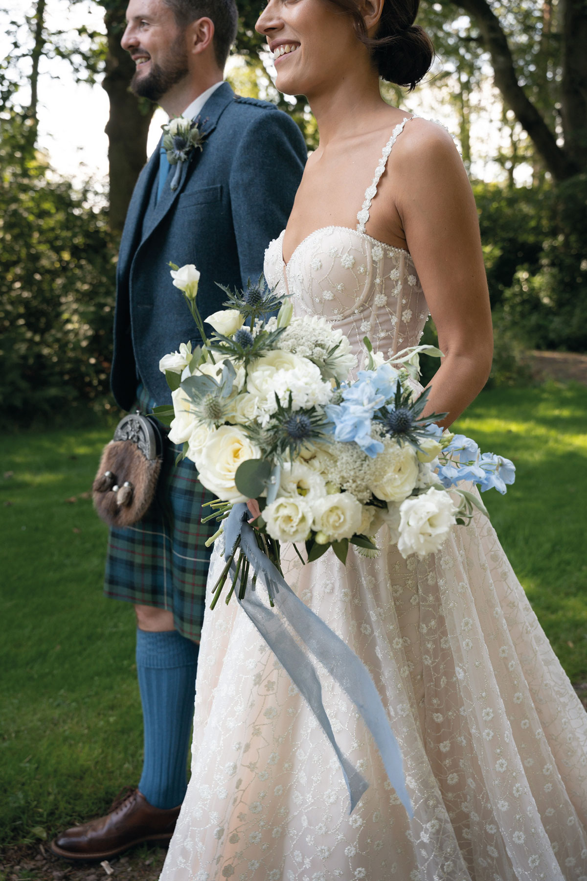 Bride in a floral embroidered wedding dress holding a bouquet of white and pale blue flowers while walking outdoors beside a groom in a blue tartan kilt suit