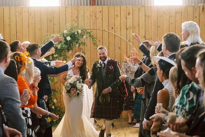 A bride and groom leaving their ceremony in a barn as the guests throw confetti over them