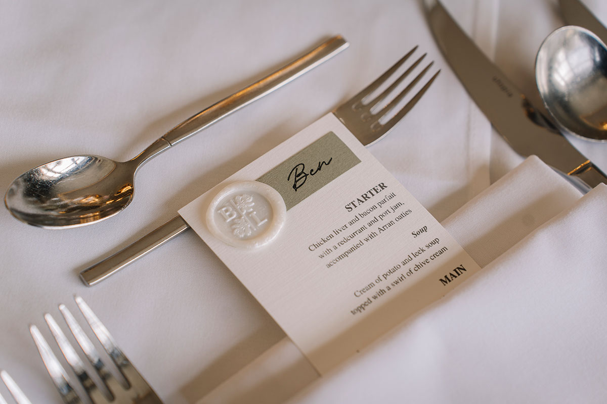 Wedding place setting with personalised menu card, wax seal detail and silver cutlery on a white linen table