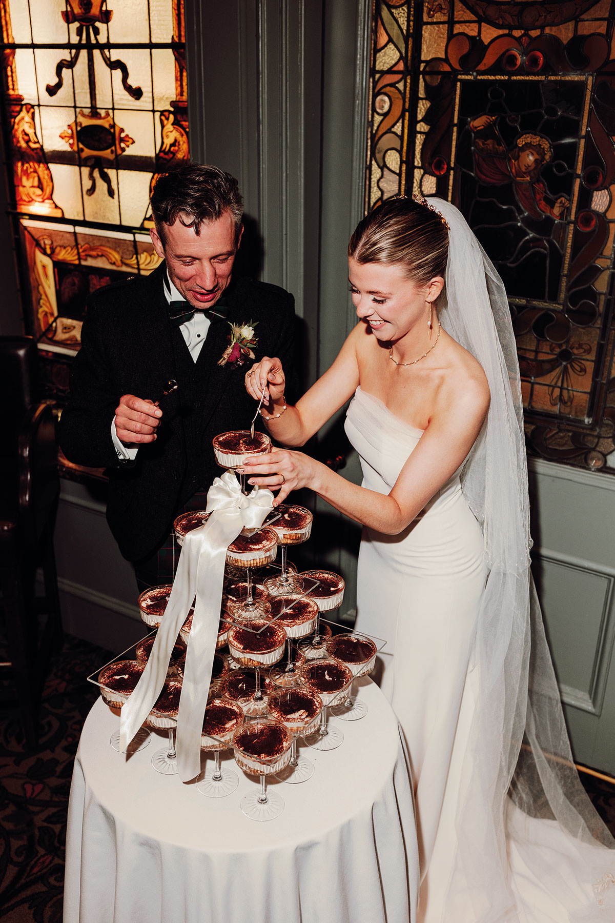 A newlywed couple smiles as they cut into a stacked tiramisu tower at their wedding reception, surrounded by stained-glass details