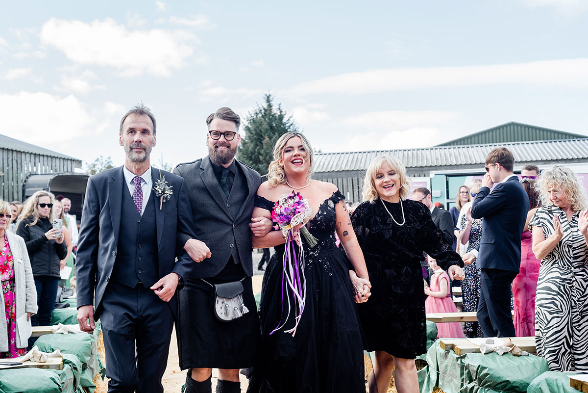 A Bride And Groom Wearing Black Walking Arm In Arm Outside With Two People, One Wearing A Black Short Dress And Another Wearing A Dark Grey Suit