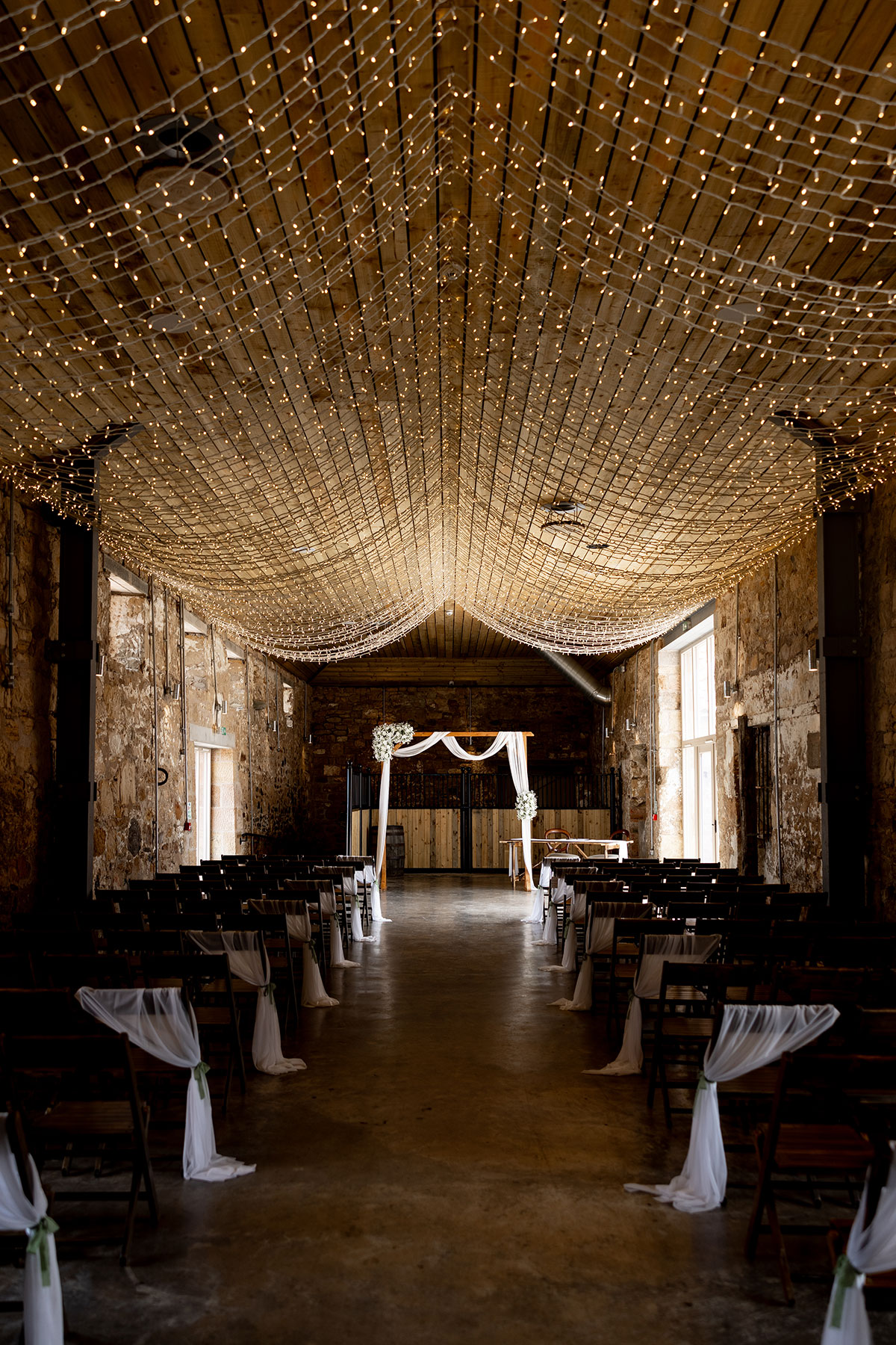 Falside Mill’s rustic ceremony space decorated with white drapes, wooden chairs and a canopy of warm string lights.