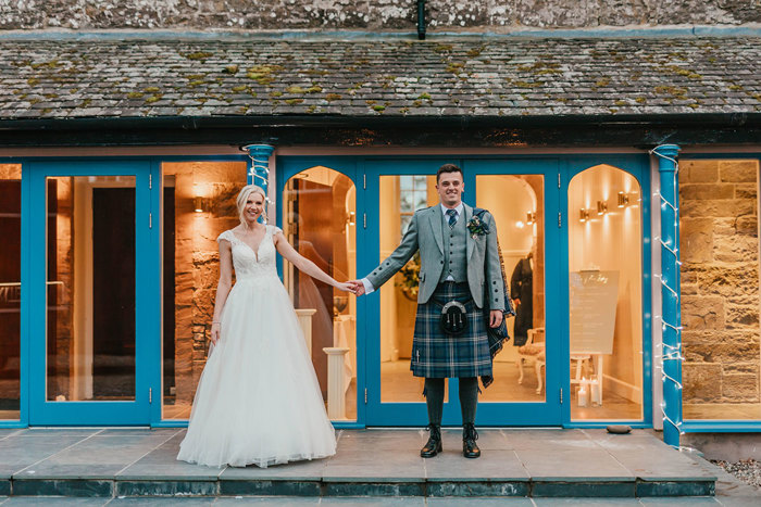 A bride in a long white dress and a groom in a blue and grey kilt face the camera holding hands standing in front of glass doors with blue frames