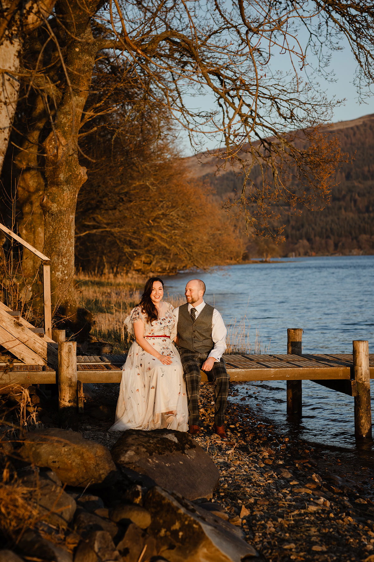 Bride and groom sitting on the dock by the loch at sunset