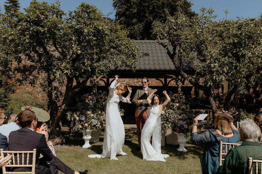 lesbian wedding ceremony outdoors in the walled garden at the Byre at Inchrya. The two brides are cheering with hands in the air.