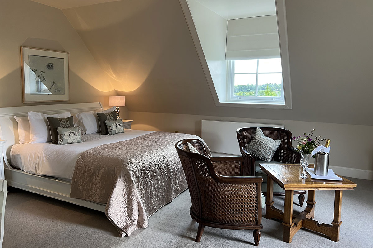 a large bed sits neatly-made on the left side of room while right side has two wooden lounge chairs and a small coffee table with champagne and glasses on it