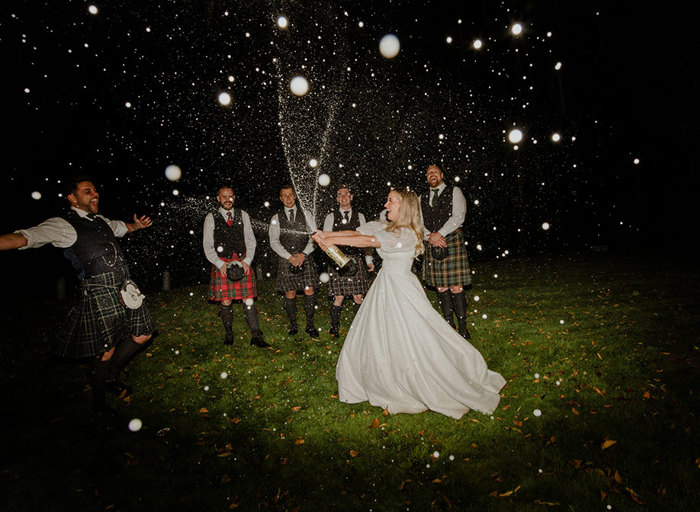 a bride points a bottle of champagne and sprays at a her groom in a kilt at nighttime as four men in kilts stand on grass in the background