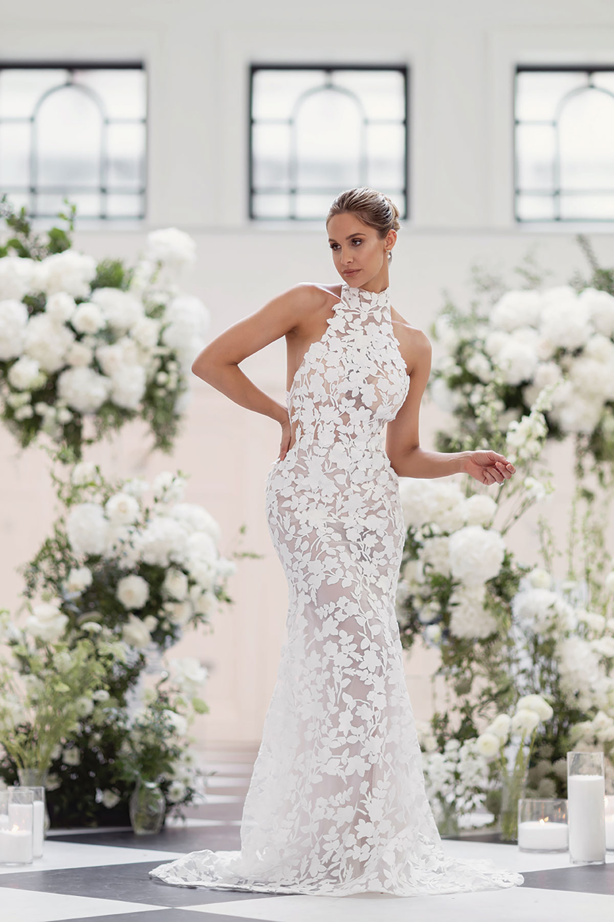Full-length view of bride in a fitted lace halterneck gown with floral details, surrounded by tall white flower arrangements